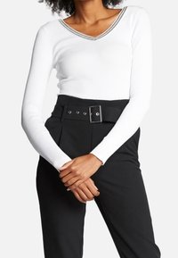 Mujer con una blusa blanca de manga larga y escote en V, metida dentro de pantalones negros de cintura alta con un cinturón ancho y hebilla rectangular.
