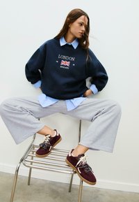 Felpa navy con la scritta "LONDON" e grafica della bandiera britannica, camicia color azzurro chiaro con colletto, pantaloni grigi a gamba larga e sneakers bordeaux con lacci.