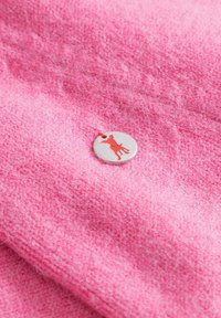 Roze textiel met een gestructureerd oppervlak en een cirkelvormig zilveren label met een rode silhouet van een hond. Het oppervlak lijkt zacht en pluizig.