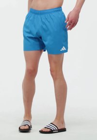 Blaue Sportshorts mit elastischem Bund und Seitentaschen; versehen mit dem weißen Adidas-Logo auf der linken Seite. Getragen mit schwarz-weißen Badeschlappen.