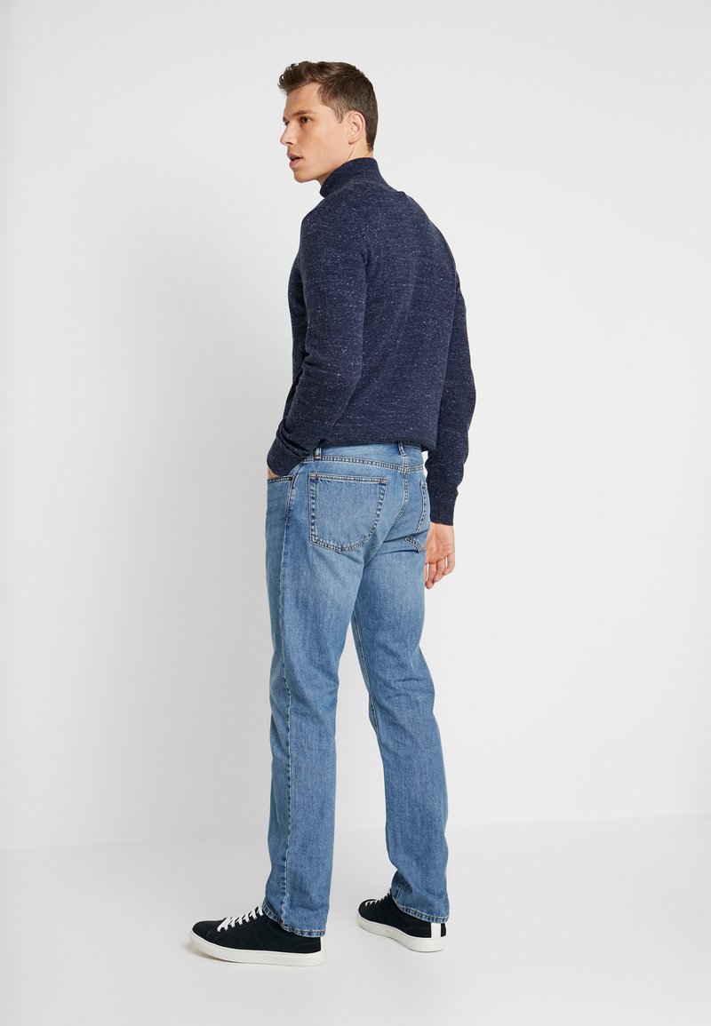 Marineblauer gestrickter Rollkragenpullover, hellblaue Jeans und schwarze Sneakers. Das Outfit zeichnet sich durch ein strukturiertes Strickmuster und eine lässige Passform aus.