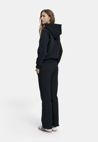 Mustahuppari, jossa on kiristettävä huppu ja ribbireunat, yhdistettyinä samanvärisiin collegehousuihin. Malli seisoo selkä kameraan päin, jalassaan valkoiset lenkkarit.