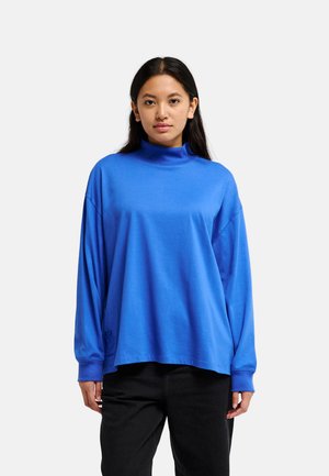 Femme aux longs cheveux foncés portant un sweat à col roulé ample bleu vif et un pantalon noir, de face contre un fond clair uni.