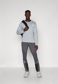 Svetlo siva majica z dolgimi rokavi z logotipom, kombinirana s temno sivimi zavihki hlač z svetlejšimi poudarki. Oblečeni v bele superge, v roki drži črno jakno.