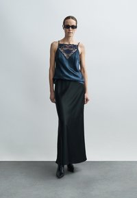 Satin-Camisole in Dunkelblau mit Spitzenbesatz am Ausschnitt, kombiniert mit einem langen schwarzen Rock. Eckige schwarze Sonnenbrille und eng anliegende Stiefel bis zum Knöchel.