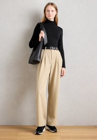 Femme portant un col roulé noir, un pantalon large beige, une ceinture noire, des baskets noires et portant un grand sac à épaule noir, debout à l'intérieur.