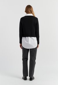 Zwarte gebreide trui met een cropped pasvorm, geribde zoom, gedragen over een witte blouse met kraag, gecombineerd met grijze rechte broek en zwarte schoenen.
