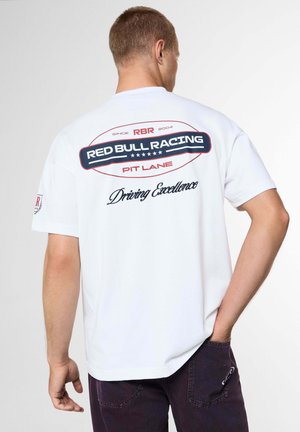Uomo con t-shirt bianca Red Bull Racing con logo blu navy e rosso e testo "Driving Excellence," in piedi con una mano in tasca, di spalle alla fotocamera.