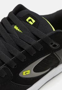 Sneaker nera con tomaia in rete e suede, lacci neri piatti, dettaglio del logo giallo neon e suola in gomma con accenti bianchi.