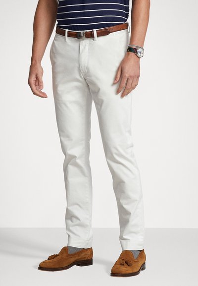 Pantalons en coton blancs ajustés, associés à des chaussures en daim marron. Une ceinture en cuir marron et une montre en argent complètent le look.