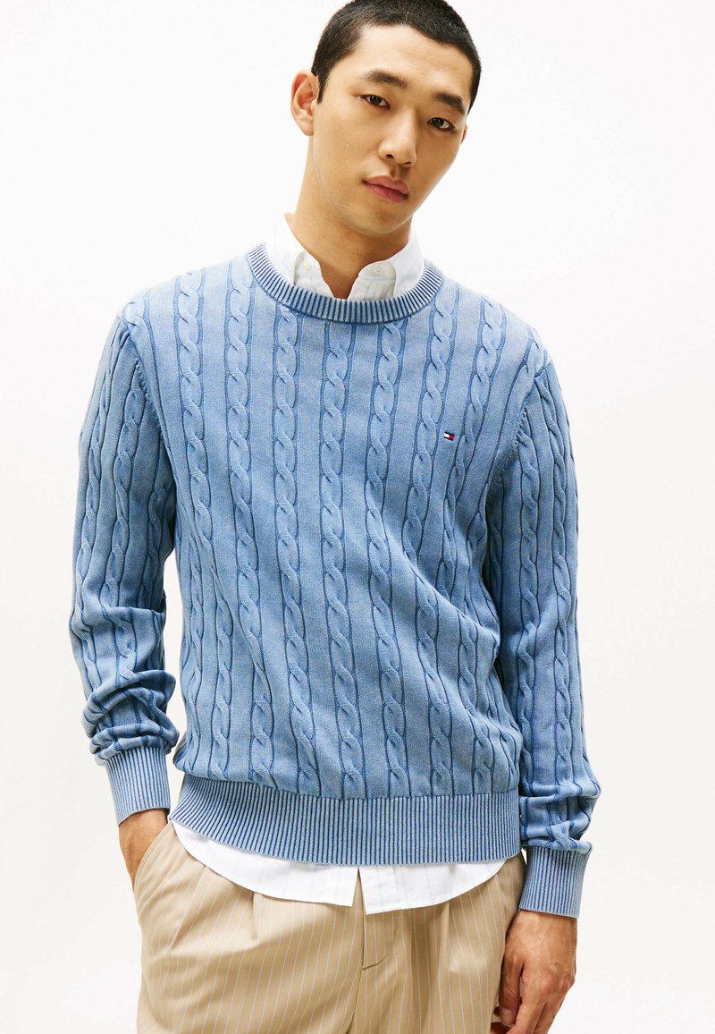 Maglione lavorato a maglia celeste con motivo a treccia, polsini e orlo a coste, indossato sopra una camicia bianca con colletto. Design casual, texture morbida.