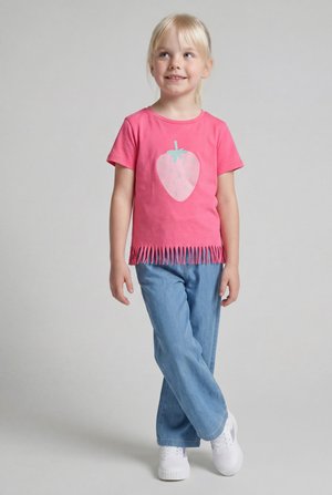 Młoda dziewczyna ubrana w różowy, z nadrukiem truskawek i frędzlami t-shirt, niebieskie dżinsy i białe trampki stoi ze skrzyżowanymi nogami, uśmiecha się i patrzy w górę.