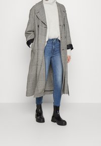 Abrigo largo en blanco y negro con estampado de pata de gallo y amplias solapas, llevado sobre un suéter de cuello alto gris claro, combinado con jeans ajustados azules y botas negras de suela gruesa.