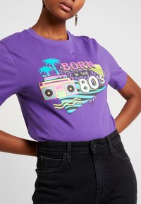 Lila bomullströja med en färgglad 80-tals grafik med palmer och en boomboxdesign. Tillsammans med högmidjade svarta jeans.