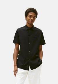 Camicia nera a maniche corte con bottoni, dal colletto ben definito, caratterizzata da un discreto logo ricamato sul petto e una superficie in tessuto liscia.