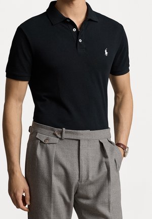 Man draagt een aansluitend zwart poloshirt met een klein wit logo, gecombineerd met hooggetailleerde grijze geplooide broek en een polshorloge.