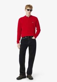 Suéter de punto rojo con cuello acanalado, jeans negros y zapatillas de deporte verde oscuro. El modelo lleva gafas de sol. Logo de Lacoste en el suéter.