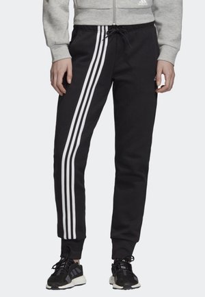 Personne portant un pantalon de survêtement Adidas noir avec trois bandes blanches sur les côtés, associé à des baskets Adidas noires et une veste zippée grise.