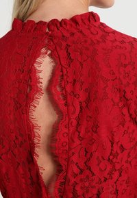 Punainen pitsimekko, jossa on kukkakuviot, korkea röyhelökaulus ja avainaukko selässä, jossa on ensiöreunat tekstuurin ja yksityiskohtien lisäämiseksi.