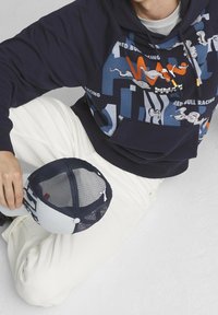 Felpa navy con vivaci grafiche che presentano il testo Red Bull Racing. Pantaloni della tuta bianchi. Cappellino da baseball in rete grigio e navy con logo.