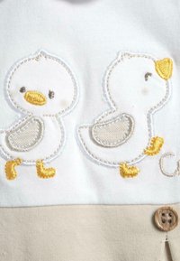 Tissu blanc avec des canards brodés en gris, jaune et blanc. Les caractéristiques des canards incluent les becs, les pattes et les détails. Accents beiges avec un bouton en bois.
