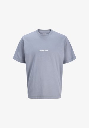 Αγόρι στέκεται χαμογελώντας, φοράει μια άνετη ανοιχτό μπλε μπλούζα με κείμενο, μαύρο παντελόνι και λευκά αθλητικά παπούτσια με μαύρες ρίγες σε απλό φόντο.