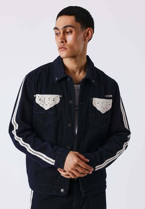 Jeune homme portant une veste en jean foncé avec des détails en crochet blanc sur les poches poitrine et les manches, regardant sur le côté les mains jointes.