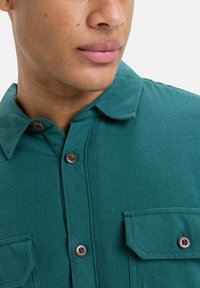 Szmaragdowa koszula zapinana na guziki z kołnierzykiem, z dwoma kieszeniami na piersi i brązowymi guzikami. Materiał ma miękką, teksturowaną powierzchnię.