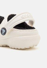 Chaussure blanche en caoutchouc avec une semelle texturée. Dotée d'un lining noir duveteux et d'une sangle ajustable blanche avec le logo de la marque sur un accent circulaire.