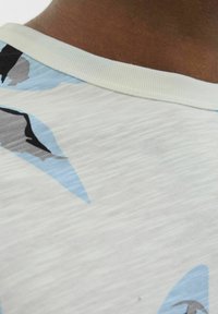 Camisa de color claro con tejido texturizado que presenta patrones abstractos en forma de hojas en azul, negro y gris, y un cuello redondo acanalado.