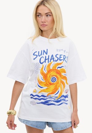 Femme blond portant un t-shirt blanc oversize avec un motif coloré de soleil et le texte "Sun Chasers,", assorti à un short en jean.
