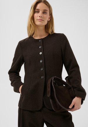 Femme aux longs cheveux blonds porte une veste boutonnée marron foncé et un pantalon assorti, tenant un sac à main en daim marron foncé avec des détails en cuir.