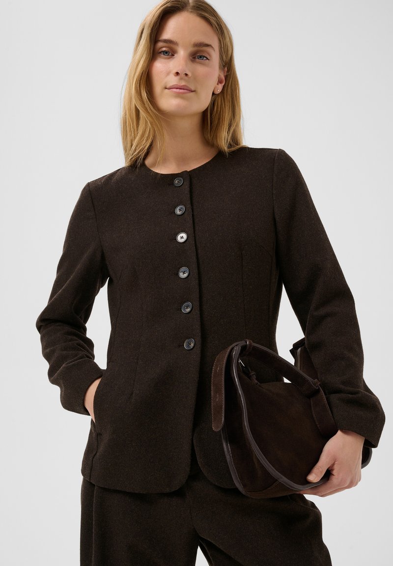 Femme aux longs cheveux blonds porte une veste boutonnée marron foncé et un pantalon assorti, tenant un sac à main en daim marron foncé avec des détails en cuir.