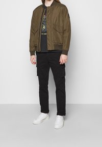 Chaqueta bomber verde oliva con cremallera frontal, puños acanalados y bolsillos laterales, combinada con pantalones cargo negros y zapatillas blancas.