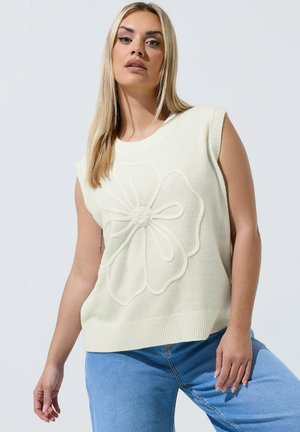Lyshåret kvinde iført ærmeløs cremefarvet sweater med stor strikket blomst og lyseblå jeans, stående mod en ensfarvet lys baggrund.