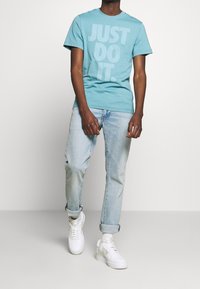 Tyrkysové tričko s velkým bílým nápisem "JUST DO IT." v kombinaci se světle modrými džíny a bílými teniskami, vystavené na neutrálním pozadí.