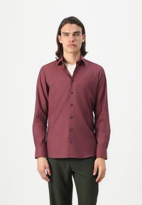 Chemise bordeaux à manches longues avec une texture subtile, fermeture à boutons à l'avant et boutons bleu marine. Portée sur une chemise crème, associée à un pantalon vert.