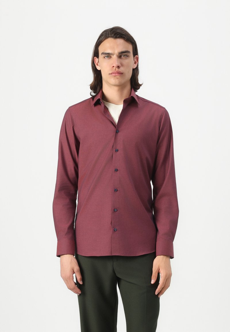 Chemise bordeaux à manches longues avec une texture subtile, fermeture à boutons à l'avant et boutons bleu marine. Portée sur une chemise crème, associée à un pantalon vert.