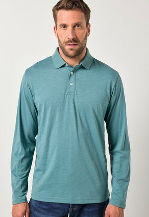 Pitkähihaisessa poolopaidassa on teal-väri ja sileä pinta, jossa on kaksipalainen nappilista ja klassinen kaulus. Paita on yhdistetty sinisiin farkkuihin.