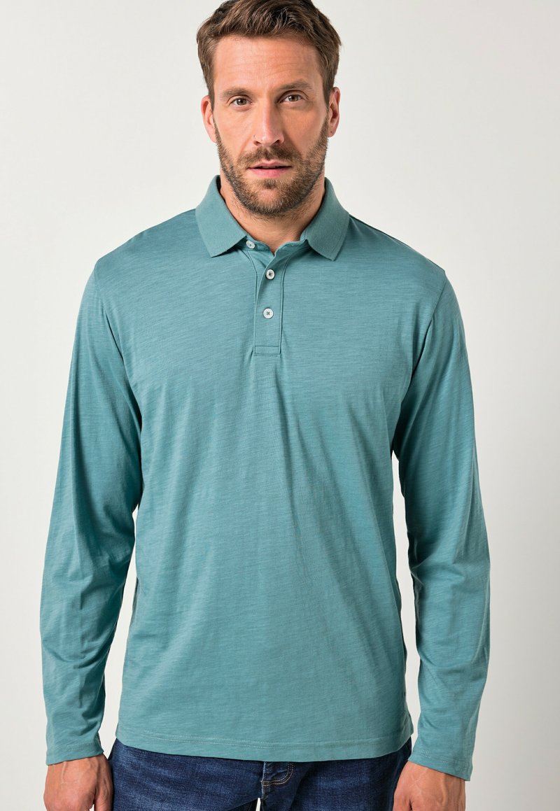 Polo a maniche lunghe in tessuto teal con una texture liscia, colletto classico e patta con due bottoni. Indossato con jeans blu.