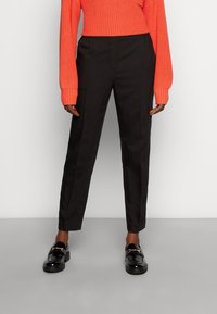 Pantalones negros de corte slim, con frente plano y bolsillos laterales, combinados con un suéter naranja texturizado y unos mocasines negros brillantes.