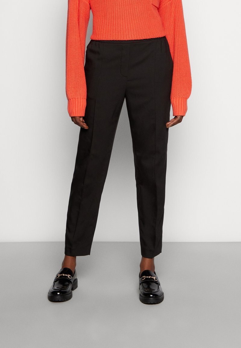 Pantalones negros de corte slim, con frente plano y bolsillos laterales, combinados con un suéter naranja texturizado y unos mocasines negros brillantes.
