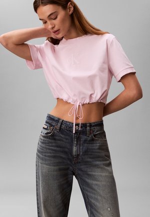 Frau trägt ein hellrosa, bauchfreies T-Shirt mit Kordelzug und hoch taillierte, ausgewaschene schwarze Jeans, posiert mit einer Hand hinter dem Kopf.