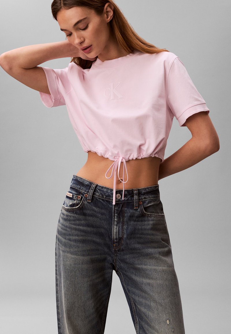 Femme portant un t-shirt court à cordon de serrage rose clair et un jean noir délavé taille haute, posant avec une main derrière la tête.