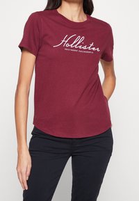 Camiseta de algodón burdeos con el logo impreso en blanco de "Hollister" y el texto "Southern California", con mangas cortas y un dobladillo redondeado.