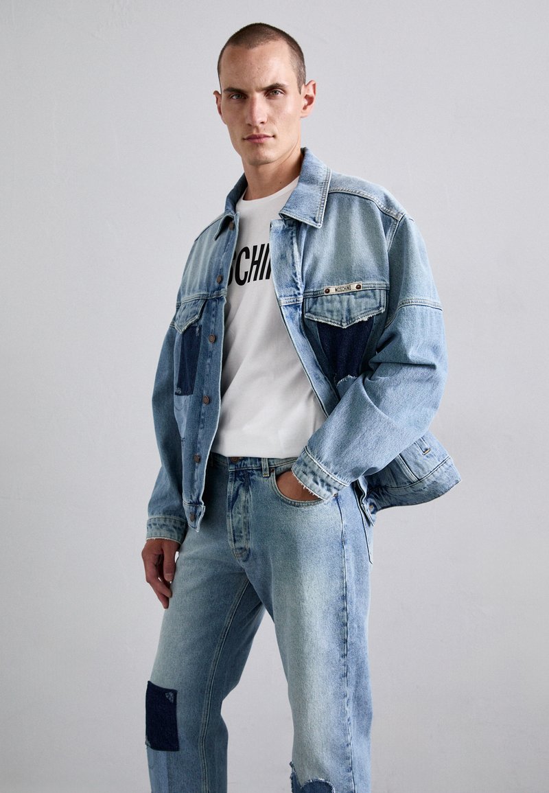 Denim jacket in light blue with patchwork accents, featuring button closure and chest pockets. Paired with light blue jeans, both distressed.