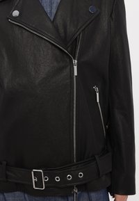 Veste en cuir noire avec fermeture éclair diagonale à l'avant, garnitures argentées, poche zippée et ceinture ajustable à la taille sur des vêtements bleus.