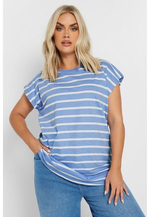 Donna bionda che indossa una maglietta a maniche corte a righe orizzontali blu e bianche e dei jeans blu, posando con una mano in tasca.