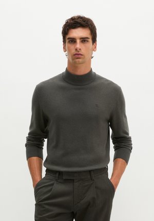 Man draagt een donkergrijze coltrui en bijpassende donkere broek, staat rechtop met handen in de zakken tegen een effen achtergrond.