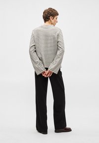 Camisola de mangas longas cinza e branca às riscas com mangas largas, combinada com calças largas pretas e sapatos escuros, vista de costas.